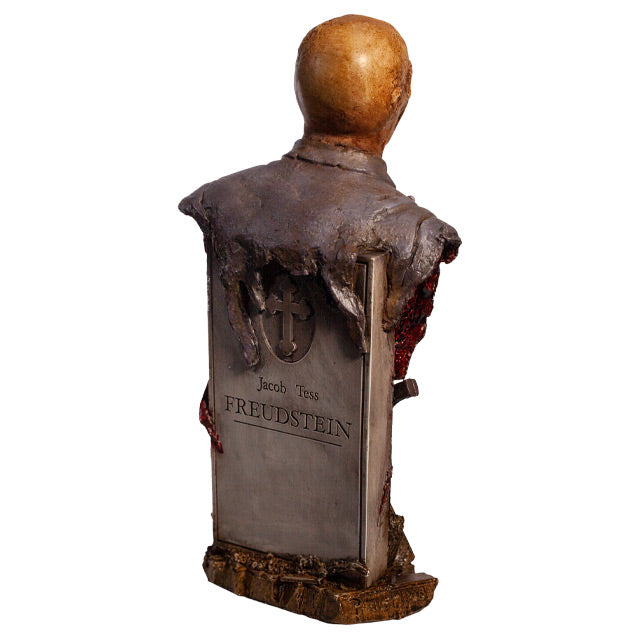 Zombie bust, back right side view, head, shoulders and chest. Orange-hued shriveled flesh, wearing filthy and gory blue doctor's coat. Gore coming from bottom of torso. Gray tombstone in back, cross in circle at top, text reads Jacob Tess Freudstein, set on wood look base.