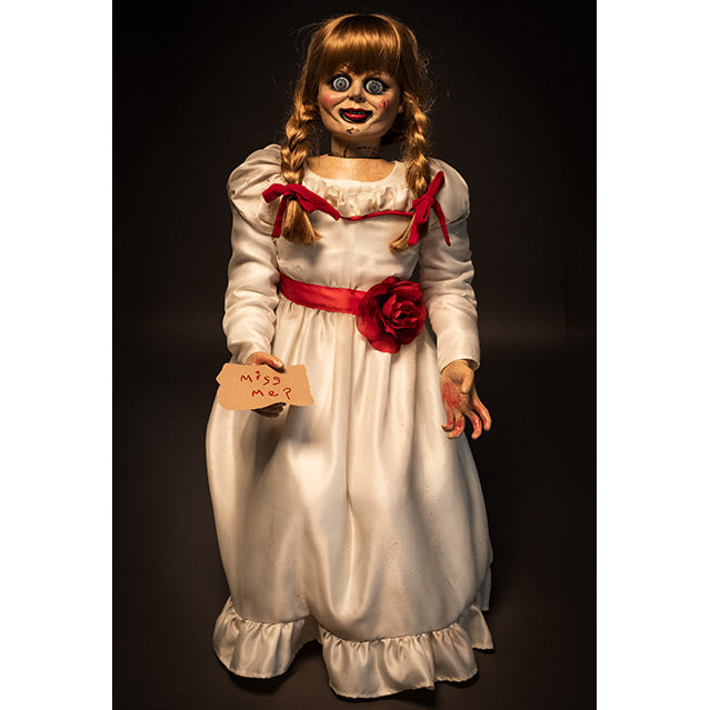 Doll, front view, black background. Large blue eyes, distressed finish on face. Blond hair with bangs, two braids tied with red ribbon. White, floor-length dress with red trim at chest and red belt with rose at waist. Holding card in right hand, red handwritten text reads Miss me? Left hand has blood on palm and fingers.