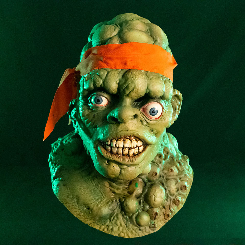Mask, head, neck and upper chest. Front view. Green lumpy blistered flesh, bald with bright orange headband tied around forehead. Misaligned blue eyes, crooked nose. Lips open showing large dirty teeth. Bumpy neck and chest with sores.  Green background.