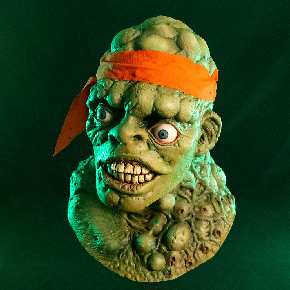 Mask, head, neck and upper chest. Left view. Green lumpy blistered flesh, bald with bright orange headband tied around forehead. Misaligned blue eyes, crooked nose. Lips open showing large dirty teeth. Bumpy neck and chest with sores.  Green background.