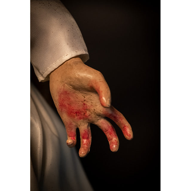Close up of doll left hand, black background. White dress, hand has blood on palm and fingers