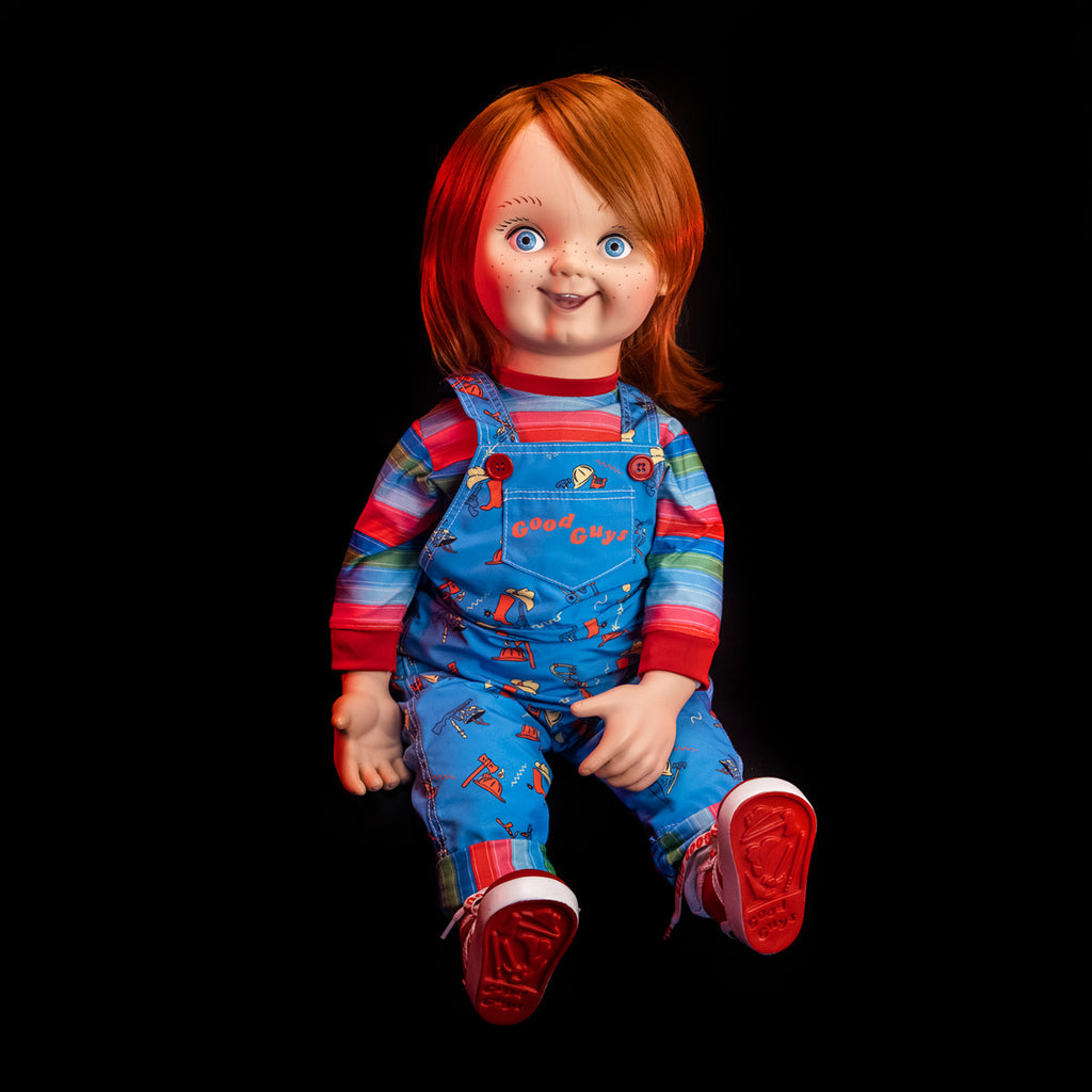 Plush body Good Guy doll sitting. Red hair, blue eyes, freckles, cleft chin. Wearing red, white, green and blue striped shirt under blue overalls, red buttons, red printed text reads Good Guys on front pocket, red and white sneakers.