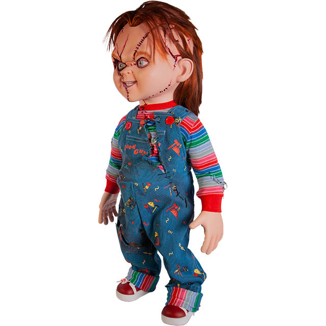 Seed of Chucky doll, left side view. Scarred Chucky, red hair, blue eyes, scarred and stitched face. Wearing a red, white, blue and green striped shirt, ripped blue overalls with red buttons, red Good Guys printed on pocket, red and white sneakers.
