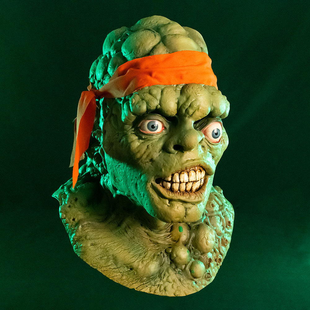 Mask, head, neck and upper chest. Right view. Green lumpy blistered flesh, bald with bright orange headband tied around forehead. Misaligned blue eyes, crooked nose. Lips open showing large dirty teeth. Bumpy neck and chest with sores. Green background.