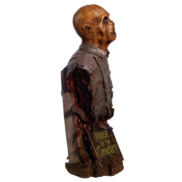 Zombie bust, left side view, head, shoulders and chest. Orange-hued shriveled flesh, wearing filthy and gory blue doctor's coat. Gore coming from bottom of torso. Plaque at bottom, gold text reads House by the Cemetary, on wood look base.