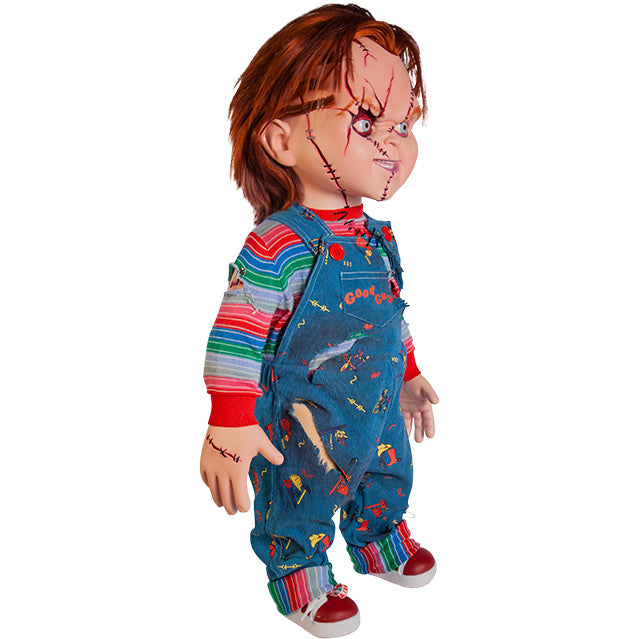 Seed of Chucky doll, right side view. Scarred Chucky, red hair, blue eyes, scarred and stitched face. Stitches on right hand. Wearing a red, white, blue and green striped shirt, ripped blue overalls with red buttons, red Good Guys printed on pocket, red and white sneakers.