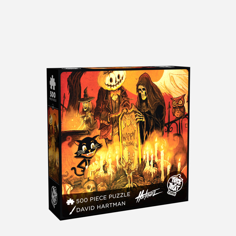 Halloween at the Cemetary jigsaw puzzle box cover. Illustration, orange, black and white cemetary scene. Skeleton in black robe and jack o' lantern creature in a suit with a bowtie standing behind gravestone with writing reading Happy Halloween, witch and owl in background, smiling cartoon-like black cat gazing at candles and skeletal hand rising out of a grave in the foreground. White text reads 500 piece puzzle, David Hartman,  White Trick or Treat Studios logo.