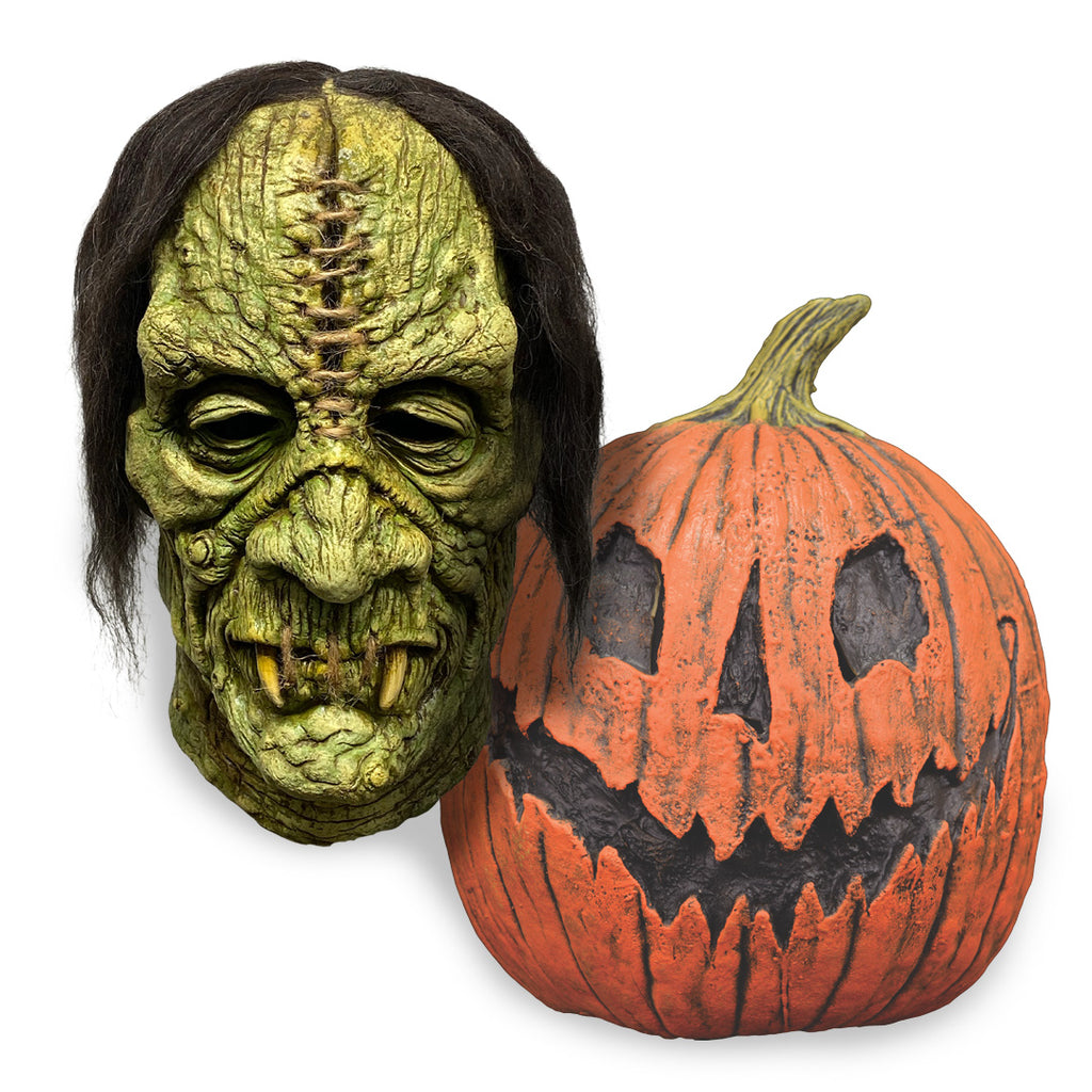 Green Halloween mask with black hair next to a carved pumpkin on a white background