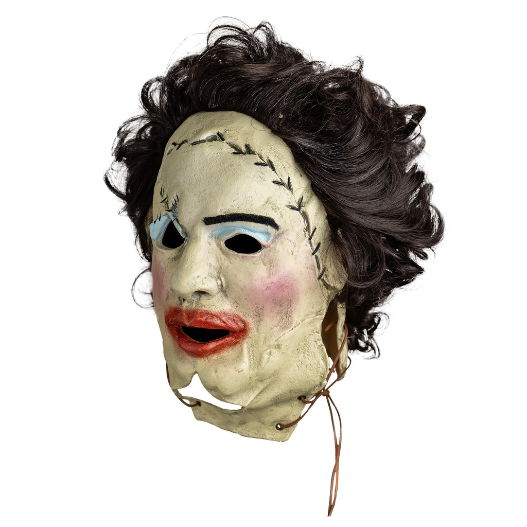 mask, left view. Woman's face, dark brown hair. stitches around upper right eye and across forehead, leather string tied at bottom sides of face. left eyebrow black, messy blue eyeshadow, pink blush on cheeks, messy overdrawn lipstick on mouth.