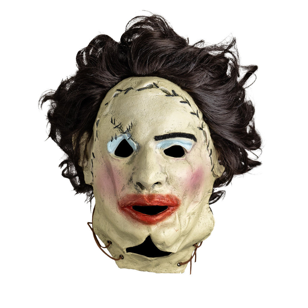 mask, front view. Woman's face, dark brown hair. stitches around upper right eye and across forehead, leather string tied at bottom sides of face. left eyebrow black, messy blue eyeshadow, pink blush on cheeks, messy overdrawn lipstick on mouth.
