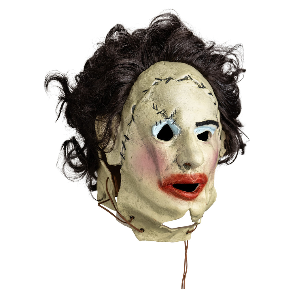 mask, right view. Woman's face, dark brown hair. stitches around upper right eye and across forehead, leather string tied at bottom sides of face. left eyebrow black, messy blue eyeshadow, pink blush on cheeks, messy overdrawn lipstick on mouth.
