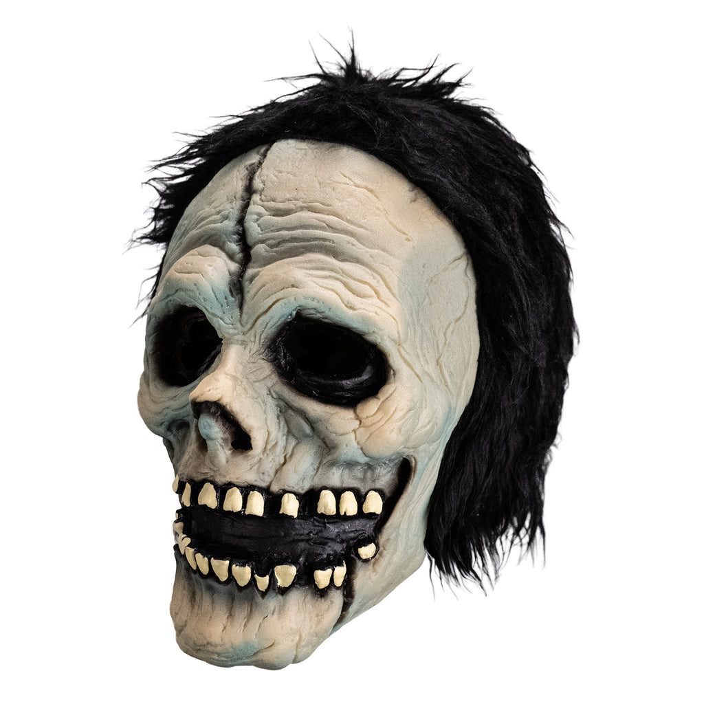 Mask, left view. White skull-like face. Black, short shaggy hair, black crack down the forehead. Black rimmed empty eyes, skull like nasal cavity. black, open grinning mouth with several missing teeth, and black crack on  left lower jaw