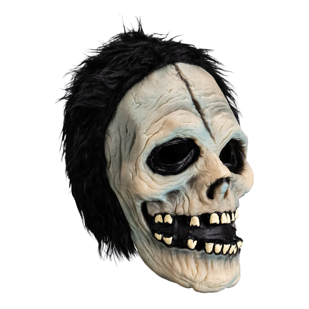 Mask, right view. White skull-like face. Black, short shaggy hair, black crack down the forehead. Black rimmed empty eyes, skull like nasal cavity. black, open grinning mouth with several missing teeth