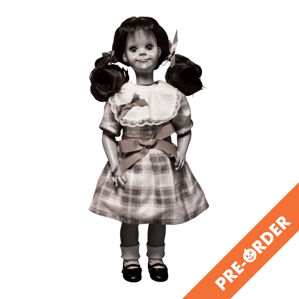 white background, orange diagonal banner at bottom right, white text reads pre-order.  Black and white illustration, Talky Tina doll wearing plaid dress, with bow at waist, hair in two ponytails with bows. Black shiny dress shoes, white socks.