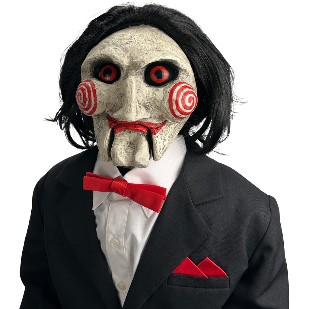 Closeup, head and shoulders view, white background. Saw Billy Puppet deluxe prop, balding with black hair, white face, black-rimmed red eyes, red spirals on cheeks, red lips on hinged ventriloquist dummy mouth. Wearing red bowtie, white collared shirt, black vest and suit coat.
