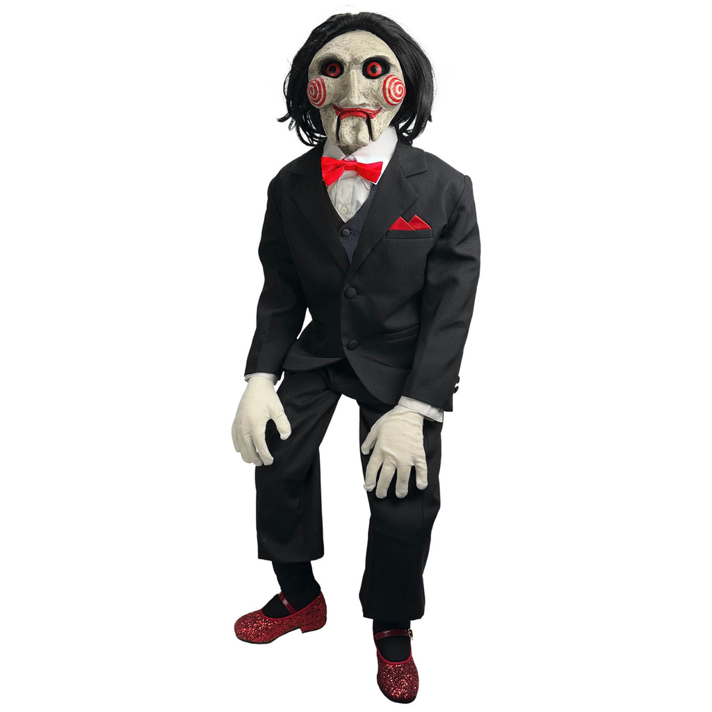 white background Saw Billy Puppet deluxe prop, balding with black hair, white face, black-rimmed red eyes, red spirals on cheeks, red lips on hinged ventriloquist dummy mouth. Wearing red bowtie, white collared shirt, black vest and suit coat, white gloves, black pants, red sparkly mary jane shoes. Standing with hands on knees.
