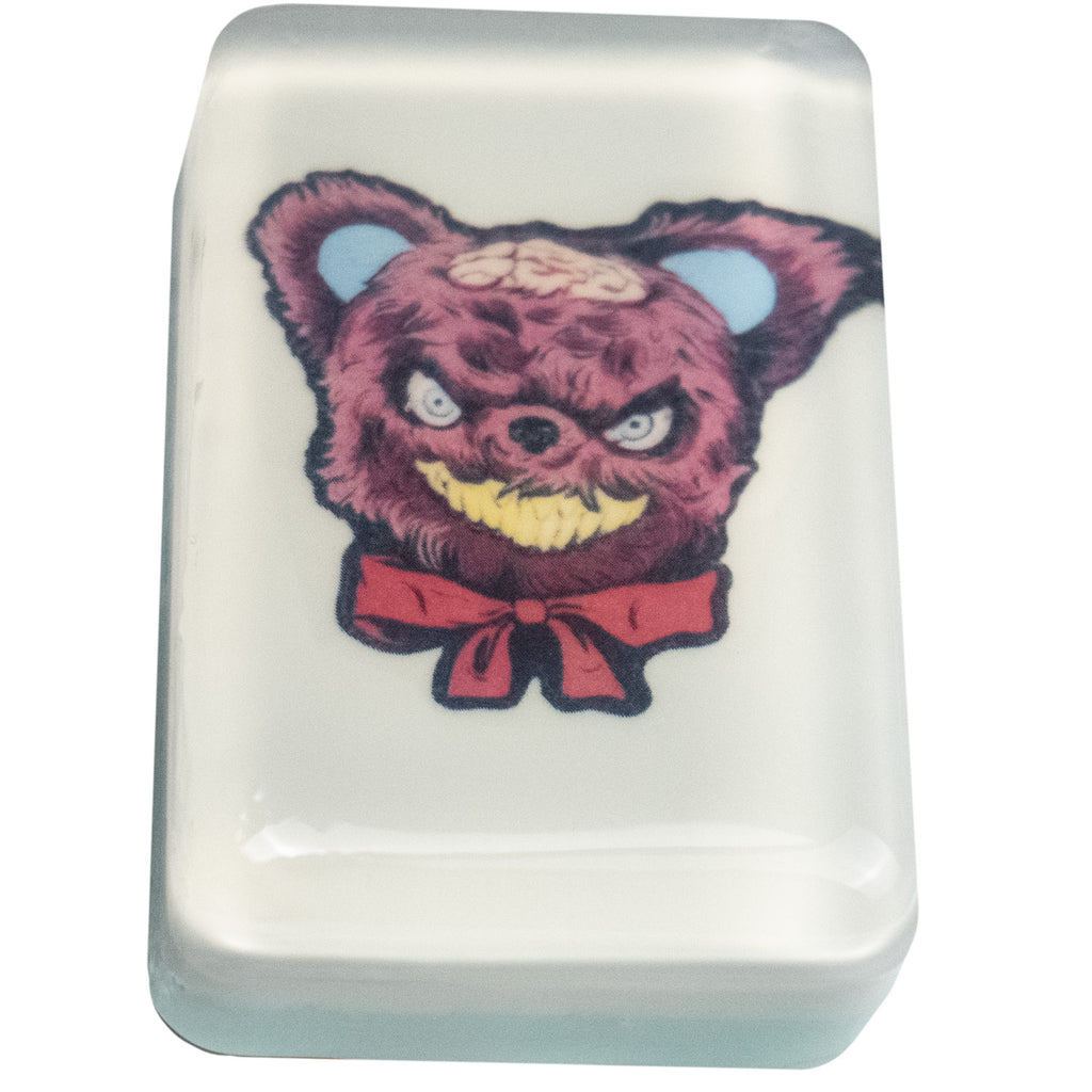 Bar soap. White, with illustration of brown Teddy bear head, blue eyes, blue inside of ears, brains exposed on scalp, menacing yellow grin, red bow tied at neck, under clear soap layer. 