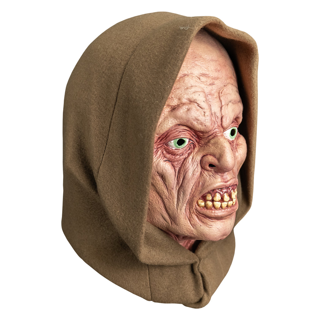 mask, right view. wearing tan hood. pink wrinkled flesh. no eyebrows, pale blue irises, lips open showing crooked yellowed teeth.