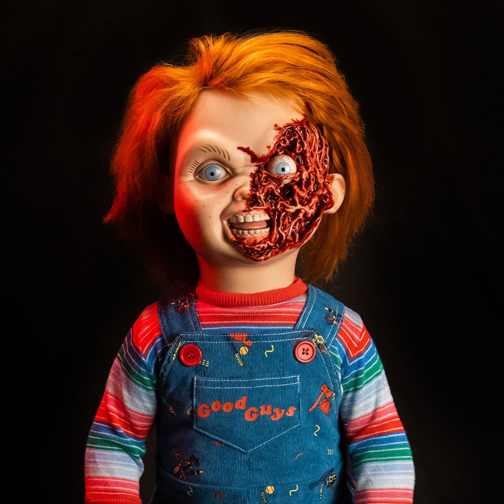 Dramatic red lighting. Ultimate Chucky Pizza Face additional head, attached to Ultimate Chucky doll. Red hair, blue eyes, freckles. Gory, fleshy left side of face, exposed eyeball. Mouth open, showing teeth and tongue. Wearing red, green, blue and white striped shirt under blue overalls. 