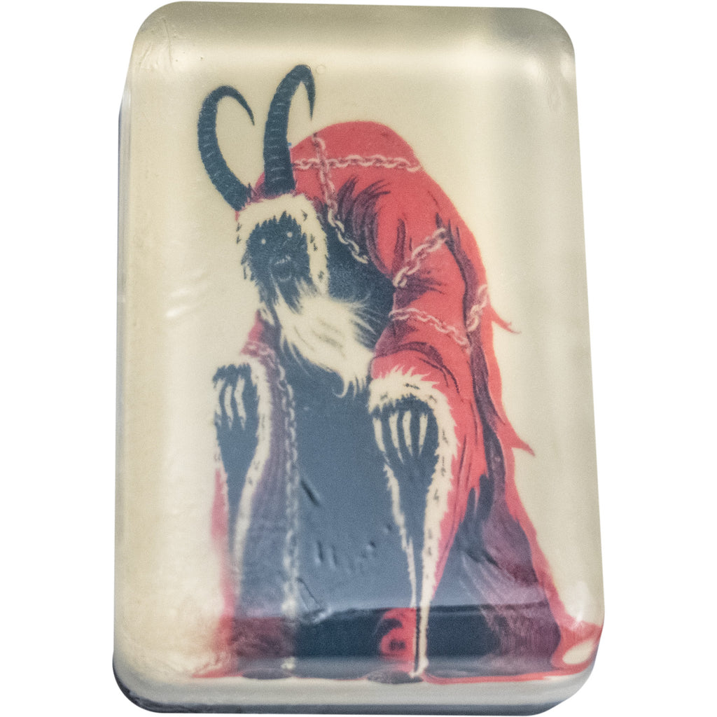 Bar soap. White, with illustration of Krampus, creature in red hooded robe with white trim, draped with chains, black face with mouth open, long curved black horns on head, clawed hands coming out of sleeves, under clear soap layer.