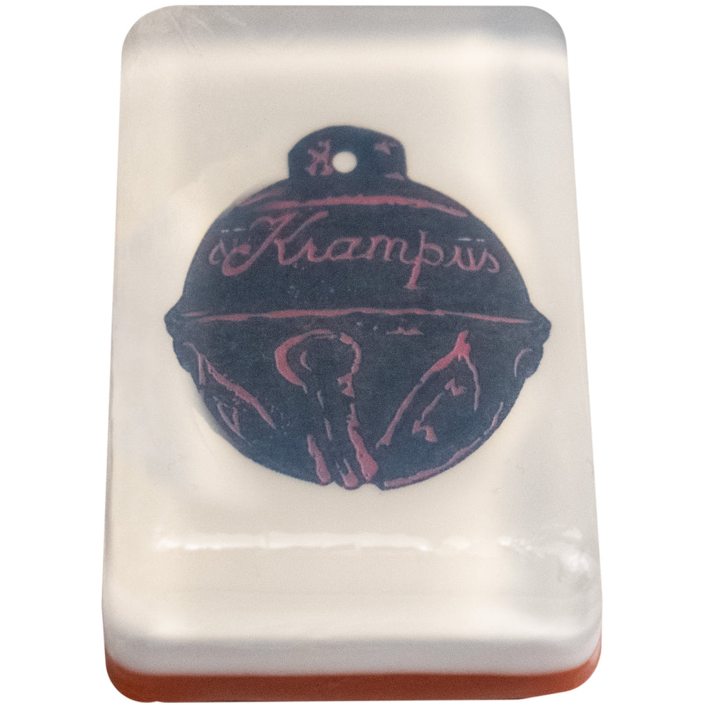 Bar soap. White, with illustration of black sleigh bell with embellishments, cursive text reads Krampus, under clear soap layer.