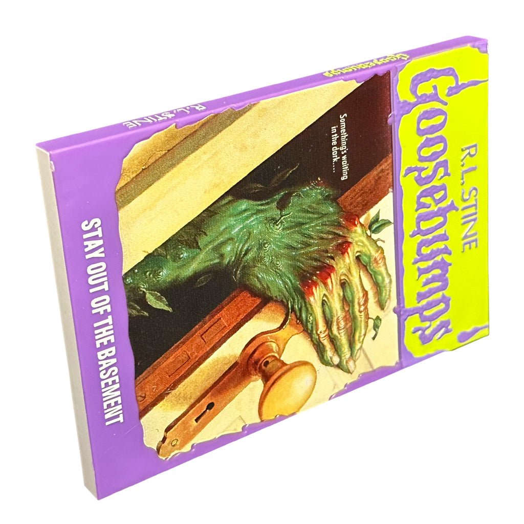 Magnet. Has the appearance of a book cover. Illustration of a door with a monster hand reaching out. Yellow banner at top, purple text reads R.L. Stine, Goosebumps. White text on illustration reads Something's waiting in the dark. Purple border at bottom, white text reads Stay out of the basement. 
