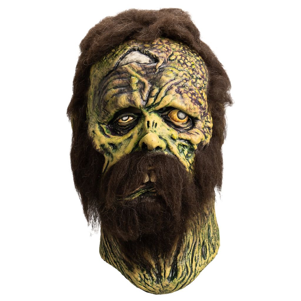 Mask, front view. Open wound on right forehead revealing skull underneath. short, matted brown hair, bushy brown sideburns and full beard with mustache. no eyebrows. Mottled, wrinkled and sagging yellow-green and brown skin. Right eye squinting almost shut. Bloodshot, open yellow left eye. small deformed nose. Crooked mouth, slightly open showing large front teeth