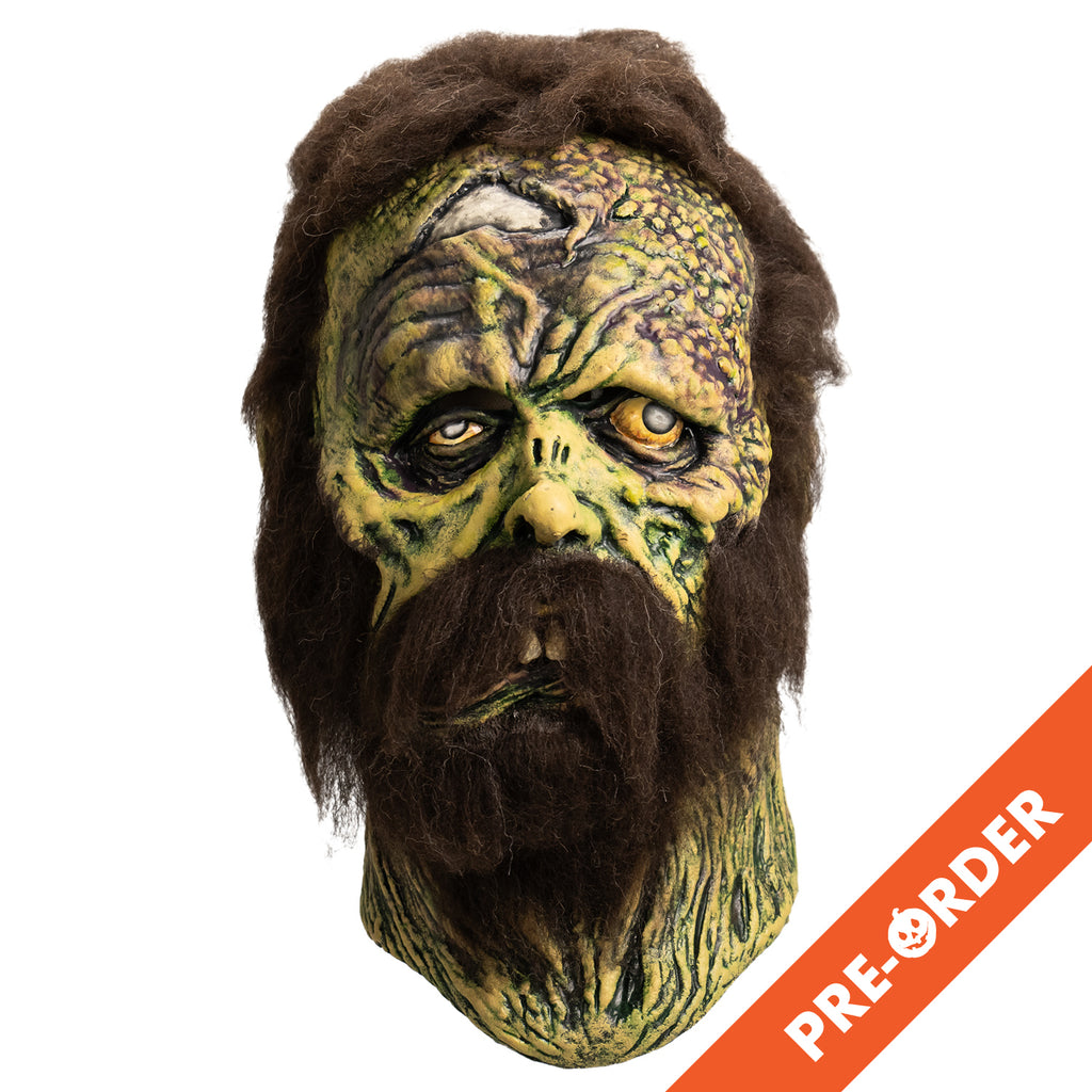 white background, orange diagonal banner bottom right, white text reads pre-order.  Mask, front view. Open wound on right forehead revealing skull underneath. short, matted brown hair, bushy brown sideburns and full beard with mustache. no eyebrows. Mottled, wrinkled and sagging yellow-green and brown skin. Right eye squinting almost shut. Bloodshot, open yellow left eye. small deformed nose. Crooked mouth, slightly open showing large front teeth