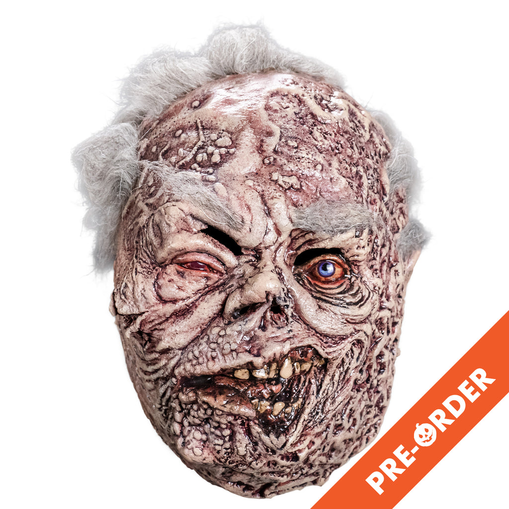 white background, orange diagonal banner bottom right, white text reads pre-order.  Mask, front view. sparse short white hair, bushy white eyebrows. Mottled, wrinkled and sagging white and brown skin. Right eye squinting almost shut and watery. Bloodshot, open blue left eye. small deformed nose. Crooked rotting mouth, sagging bottom lip, slightly open showing some crooked and dirty teeth