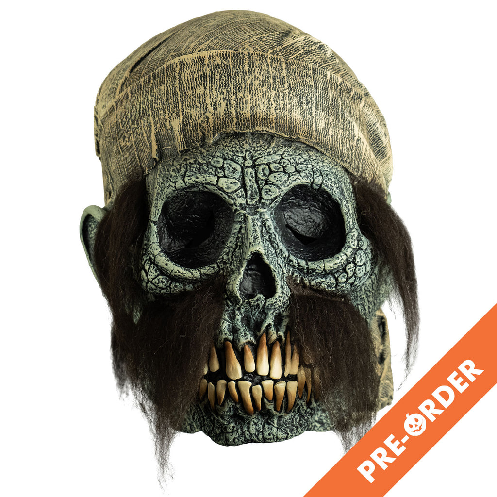 white background, orange diagonal banner, bottom right, white text reads pre-order.  mask, front view. dirty off-white scarf on head hanging down to neck. Empty eye sockets, long brown sideburns and mustache. Cracked gray skull face with ears, long yellow teeth.