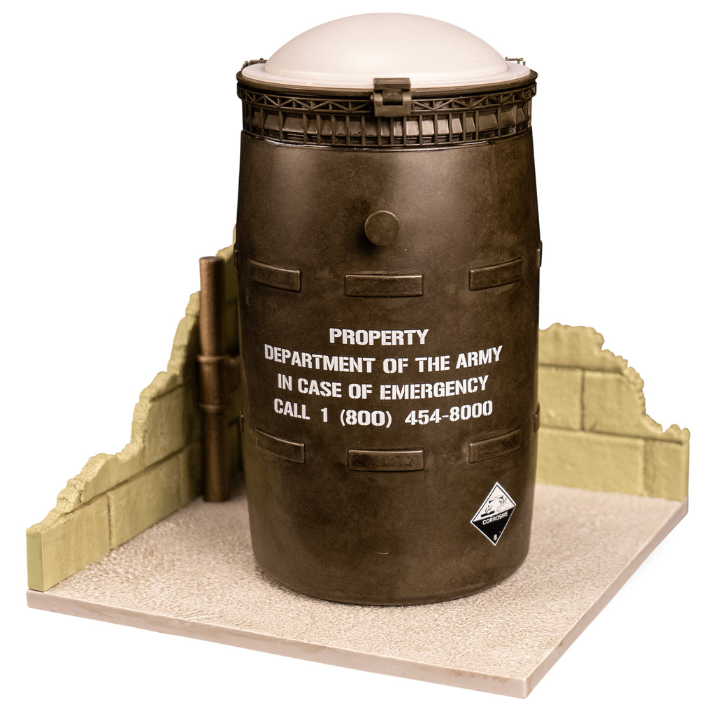Black barrel with white lid, white text reads property of the army in case of emergency call 1 800 454-8000. set on cement-look gray base with broken cement block walls on two sides.