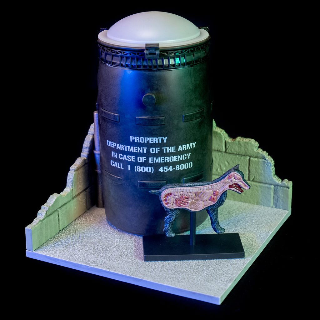 black background.  Black barrel with white lid, white text reads property of the army in case of emergency call 1 800 454-8000. anatomical figure of dog cut in half view. all set on cement-look gray base with broken cement block walls on two sides.