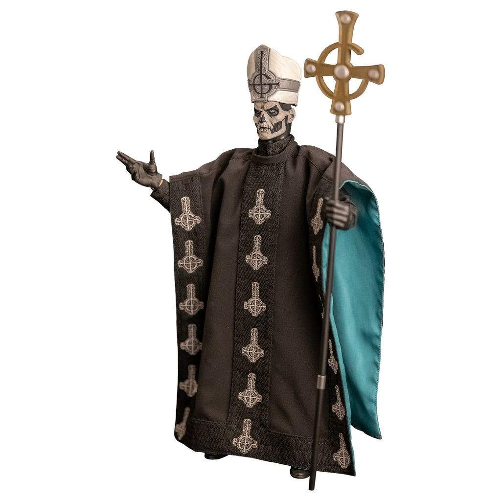 Left side view. White background. Figure and accessories. Black and white skull-like face wearing black and silver, tall hat adorned with black inverted cross with semi circle that looks like a G. wearing black floor-length robe green on the inside adorned with several white inverted crosses, with semi circle that looks like a G. black gloves and black boots. Holding black staff topped with inverted gold cross.