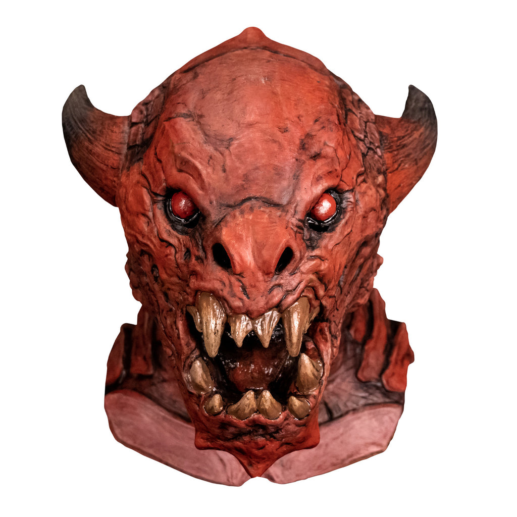 Mask, front view.  Red creature with horns on either side of head, red black-rimmed eyes, snout-like nose, open mouth showing several large yellowed sharp fangs,  wrinkled and scaly skin on face pointed chin.  Large plate-like scales on neck and chest