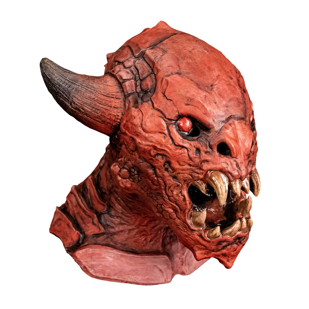 Mask, right side view. Red creature with horns on either side of head, red black-rimmed eyes, snout-like nose, open mouth showing several large yellowed sharp fangs, wrinkled and scaly skin on face pointed chin. Large plate-like scales on neck and chest