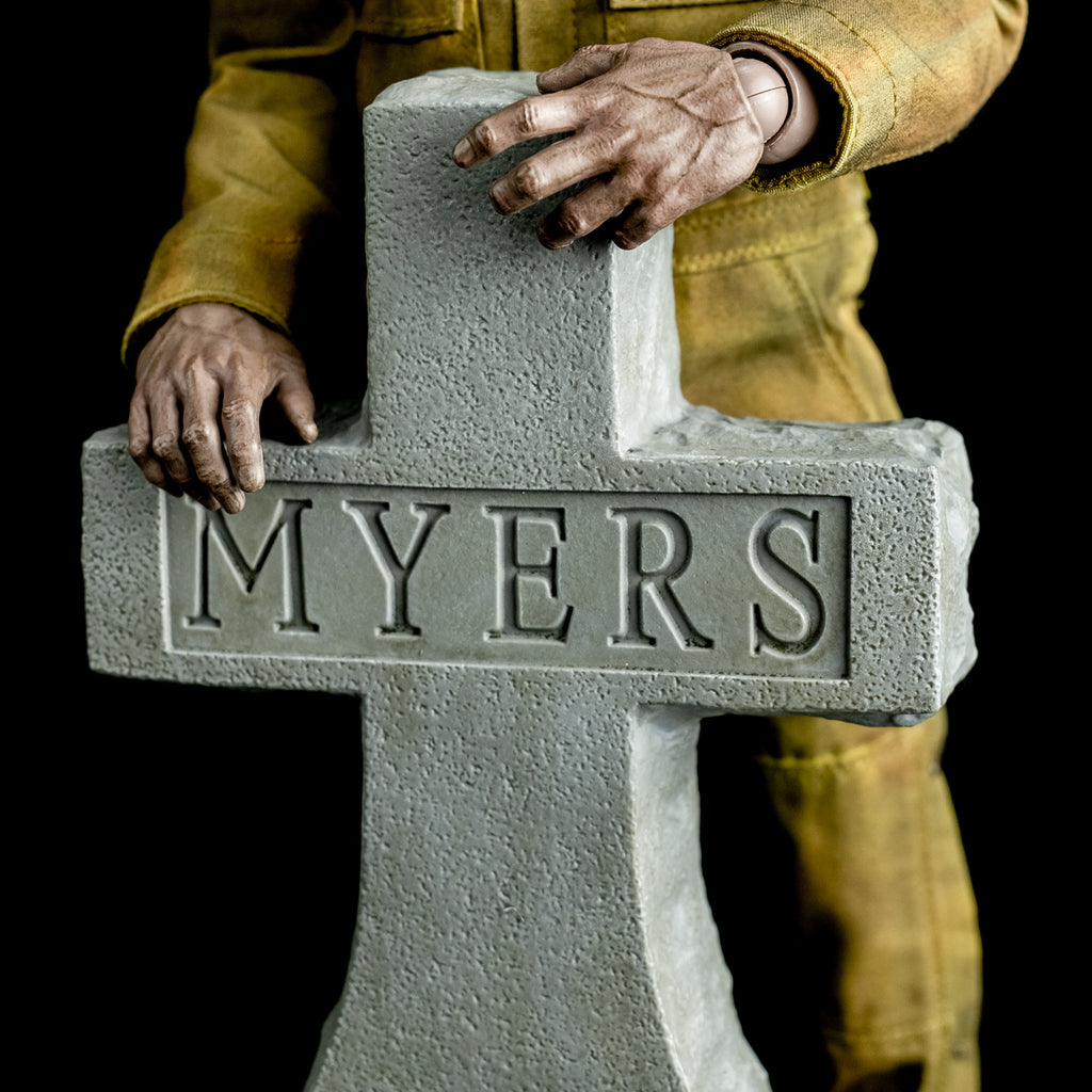 close up view, Upper half of Tombstone accessory. Gray cross tombstone, texts reads Myers. Michael Myers hands and mid-body wearing coveralls behind it.  