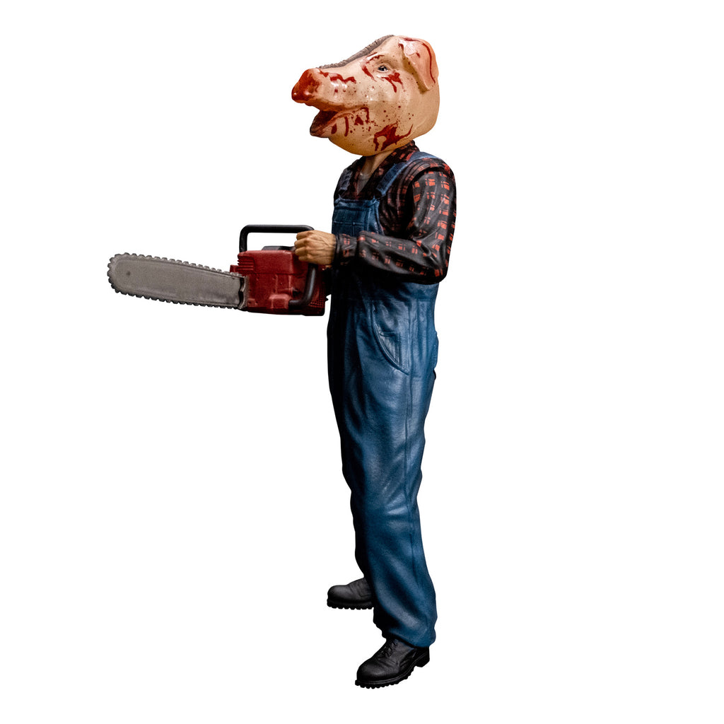 8 inch figure, right side profile view. Person with bloodied pig head mask, mouth open. Wearing flannel print shirt, blue overalls, black boots. Holding red chainsaw with both hands.