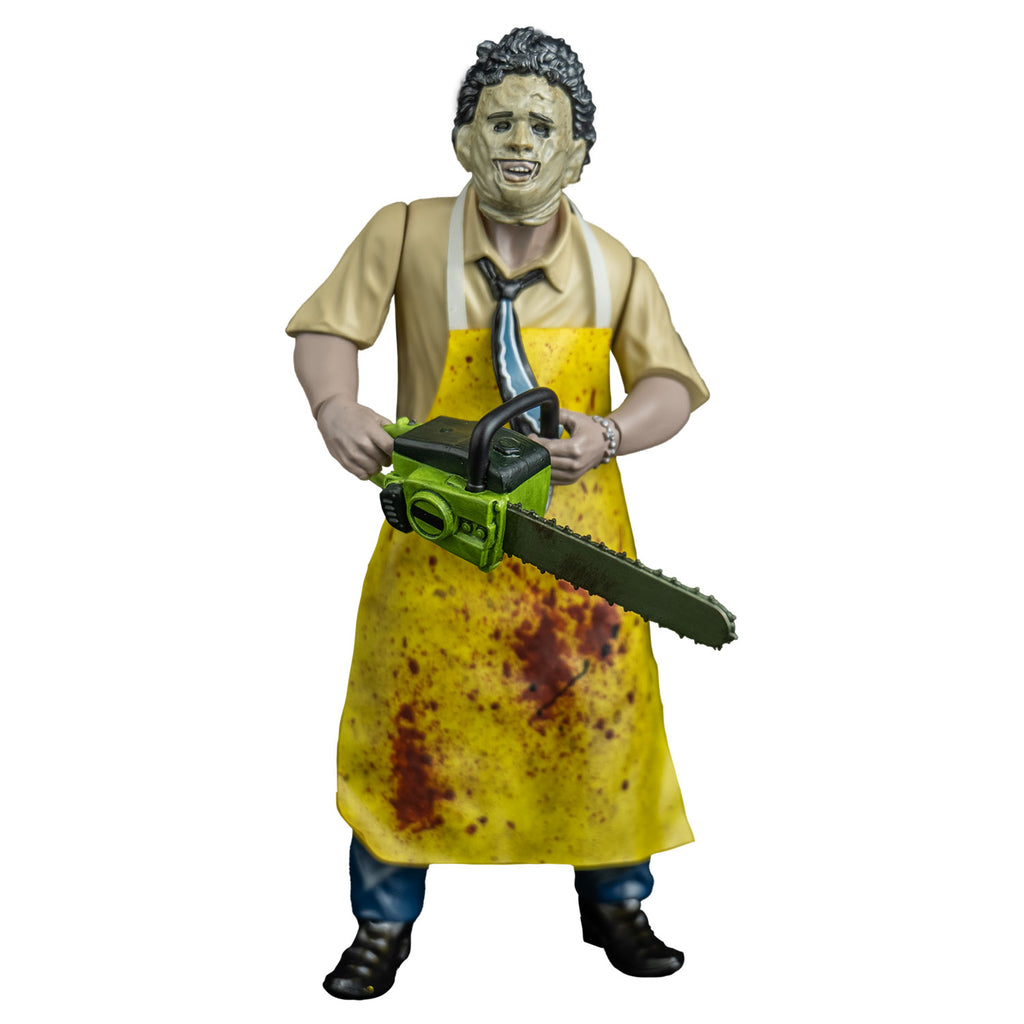 Leather face action figure, front view. wearing killing mask, short brown bushy hair, black eyebrows, skin is sewn together, stitches around forehead and ears, open space for mouth, tan button up short-sleeved shirt, black, blue and white necktie, yellow apron splattered with blood, blue pants and brown shoes.  Holding a green bodied chainsaw in both hands, at waist level