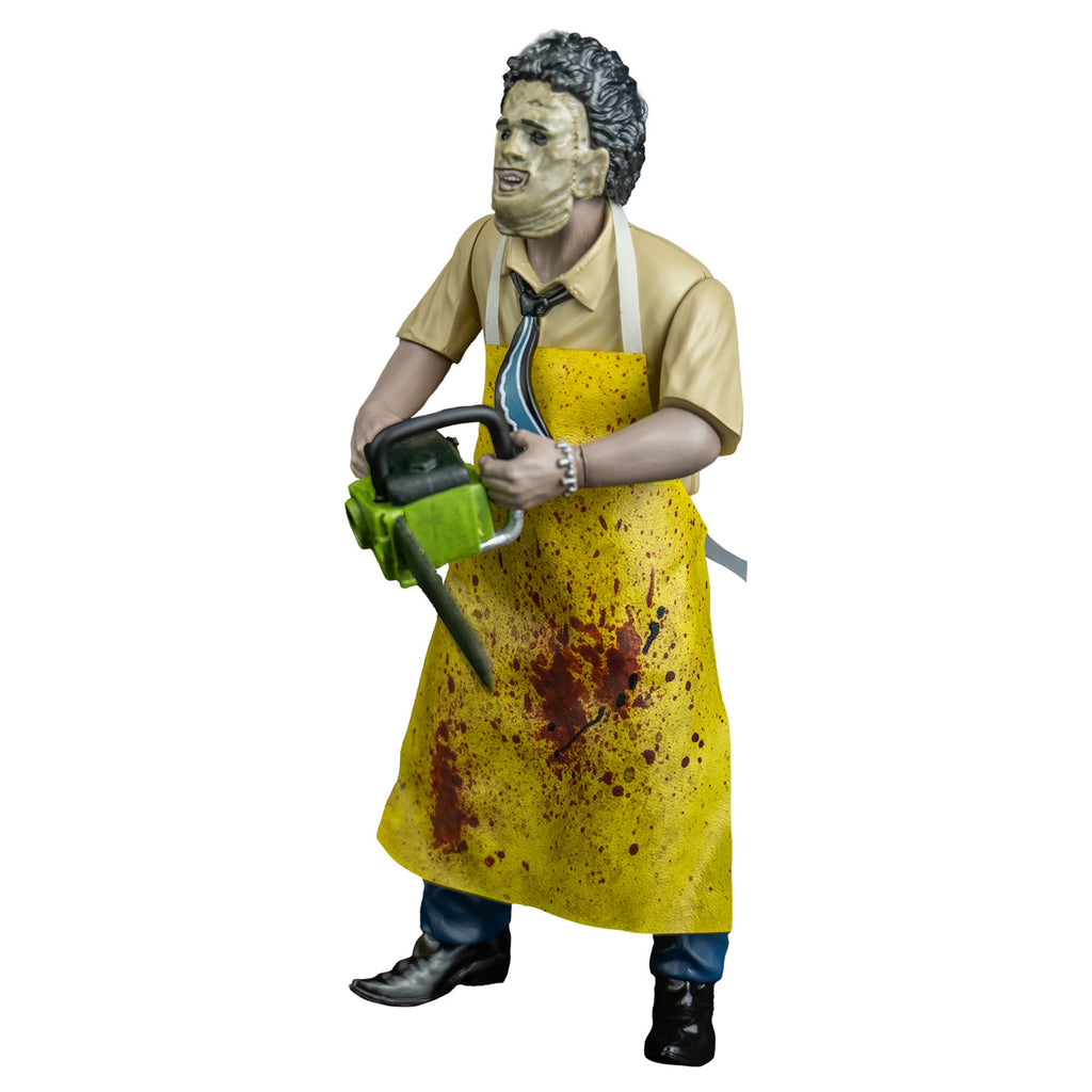 Leather face action figure, left view. wearing killing mask, short brown bushy hair, black eyebrows, skin is sewn together, stitches around forehead and ears, open space for mouth, tan button up short-sleeved shirt, black, blue and white necktie, yellow apron splattered with blood, blue pants and brown shoes. Holding a green bodied chainsaw in both hands, at waist level