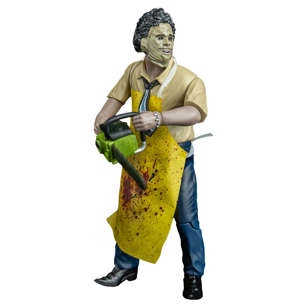 Leather face action figure, left view head turned to his left. wearing killing mask, short brown bushy hair, black eyebrows, skin is sewn together, stitches around forehead and ears, open space for mouth, tan button up short-sleeved shirt, black, blue and white necktie, yellow apron splattered with blood, blue pants and brown shoes. Holding a green bodied chainsaw in both hands, at waist level