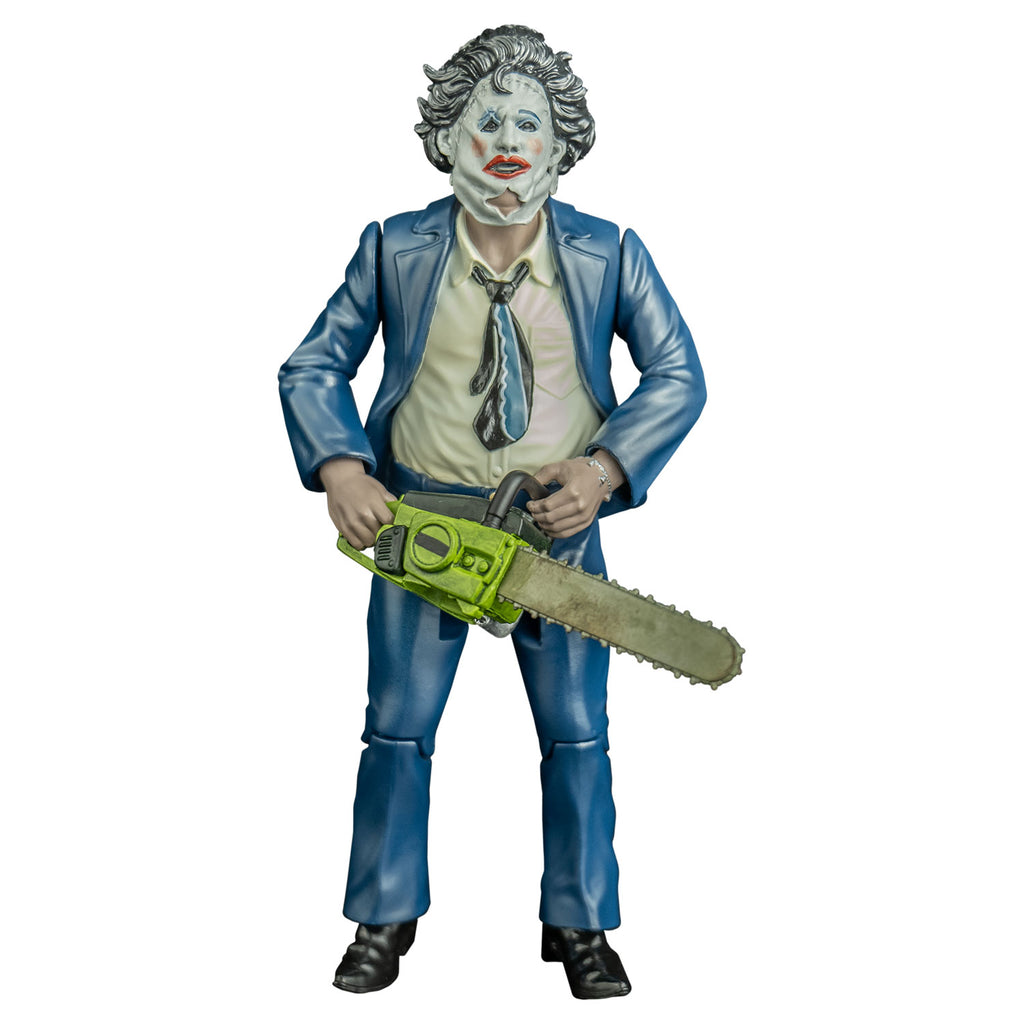 action figure, front view.  Leatherface, wearing pretty woman mask. Woman's face, dark hair. stitches around upper right eye and across forehead, leather string tied at bottom sides of face. left eyebrow black,  blue eyeshadow, pink blush on cheeks, red lipstick on mouth. Wearing blue suit coat and pants, white collared shirt, black blue and white necktie, black shoes.  Holding yellow bodied chainsaw in both hands. 