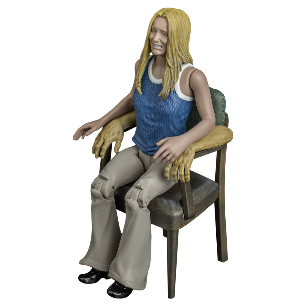 Sally figure, left side view. Blond woman, grimace on face, blue and white tank top, tan flare legged pants. Sitting in chair with human arms.