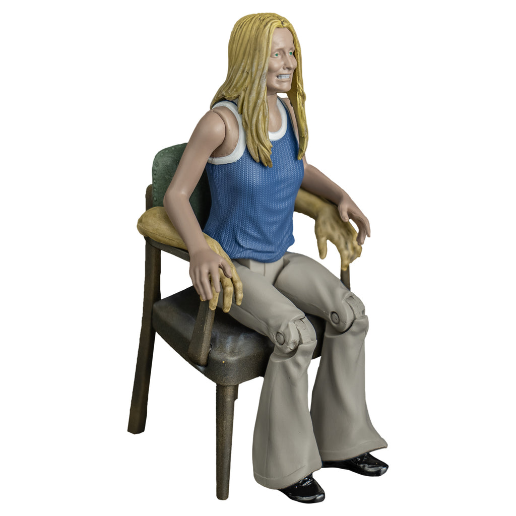 Sally figure, right side view. Blond woman, grimace on face, blue and white tank top, tan flare legged pants. Sitting in chair with human arms.