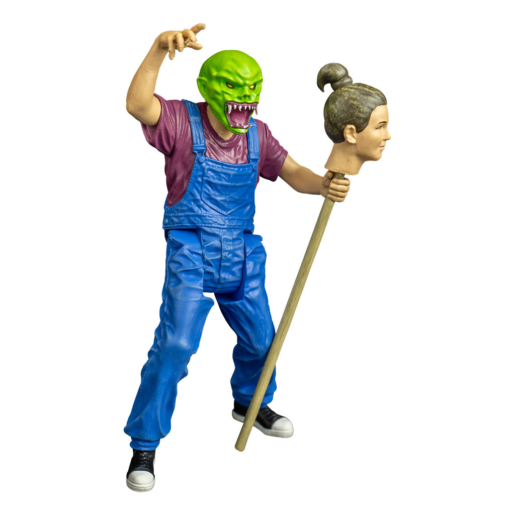 goosebumps figure, right view.  Wearing haunted mask,Bright green skin, no hair. Angry face with large ears, yellow eyes, large snarling mouth with sharp white teeth, pink tongue and gums. white drool on lower jaw and chin. Purple t-shirt, blue overalls, black and white sneakers.  Right arm bent at elbow raised over head.  left hand holding staff with person's head attached, brown hair pulled back in a bun.