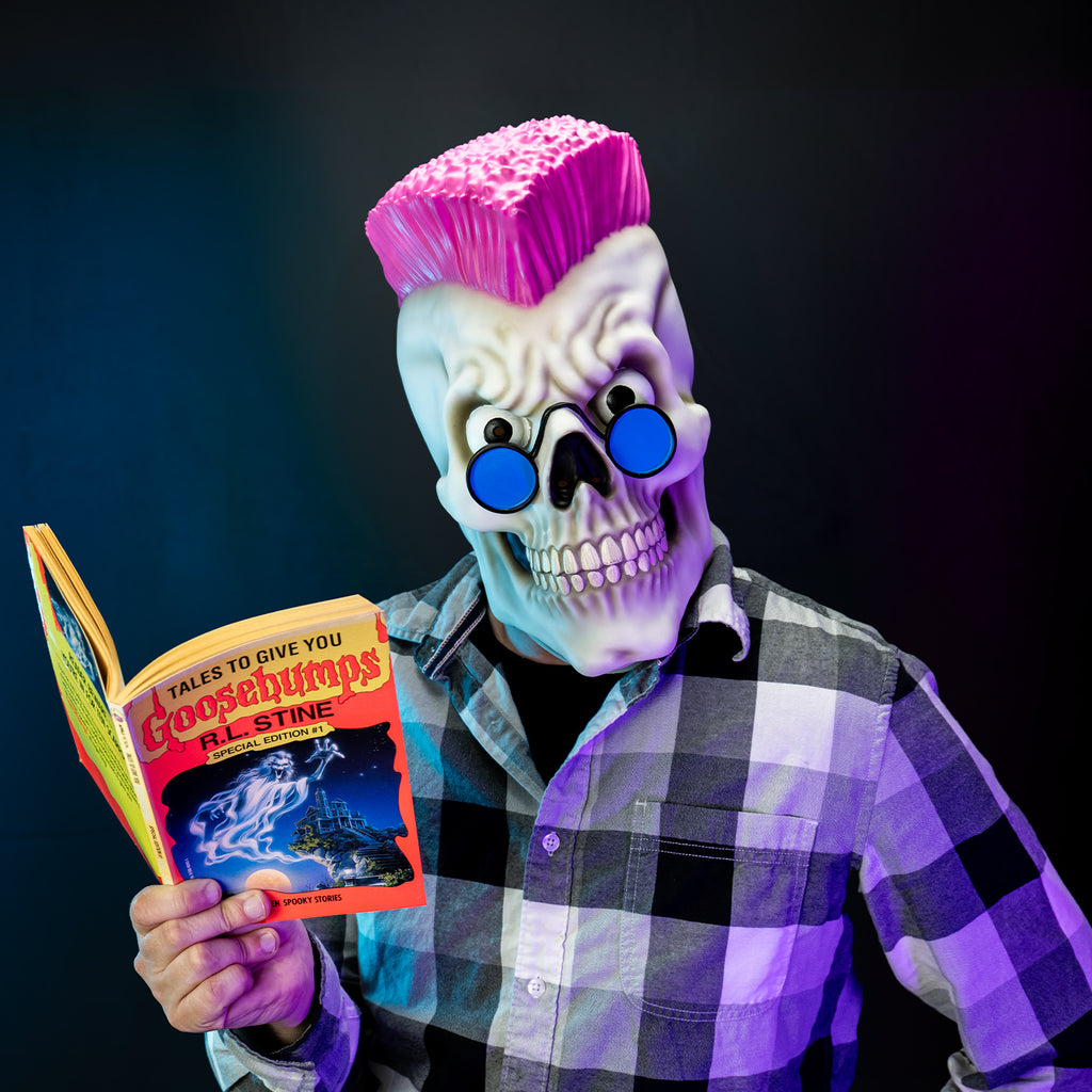 black background, Glam shot, man holding a Goosebumps book wearing black, white and gray flannel shirt and the Goosebumps curly mask, white grinning skull face, pink mohawk, large eyes, wearing blue round sunglasses.