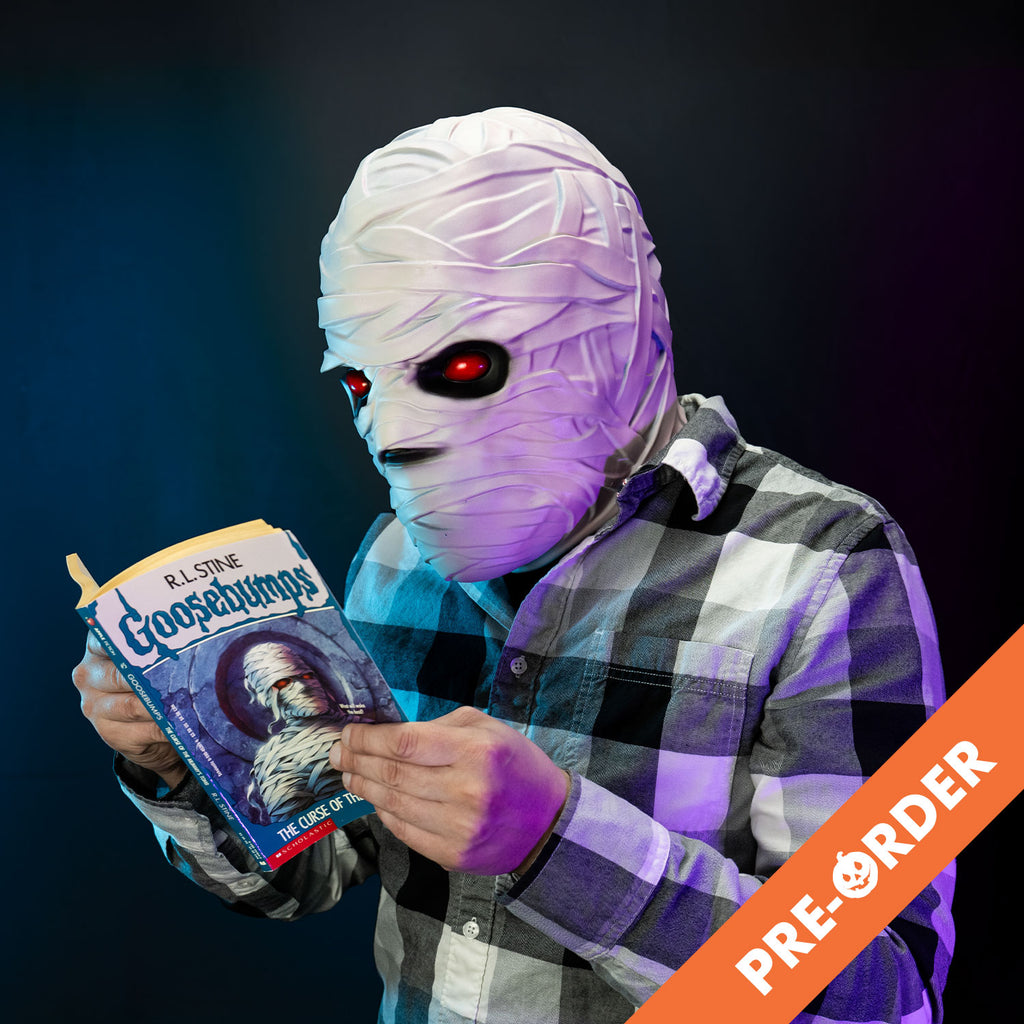 black background orange diagonal banner at bottom right, white text reads pre-order, Glam shot, man reading Goosebumps book wearing black, white and gray flannel shirt and the Goosebumps Mummy mask, white bandaged head red eyes, black eye sockets. Black on tip of nose