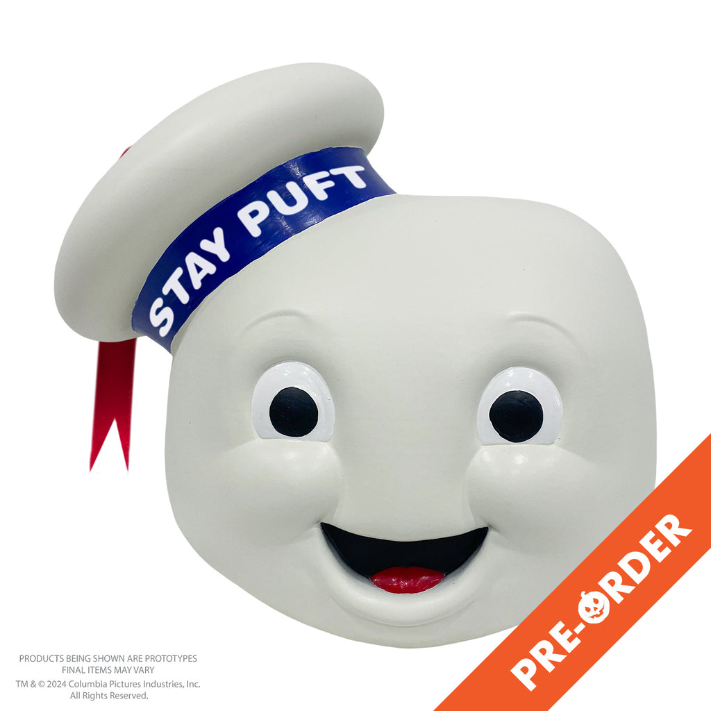 White background, orange diagonal banner at bottom right, white text reads pre-order. Mask, front view. round white head, large eyes with large black pupils, mouth open in a smile showing red tongue. White hat cocked to the side on the right side of the head, blue band with white text reads Stay Puft, red ribbon tassel hanging from the top center.