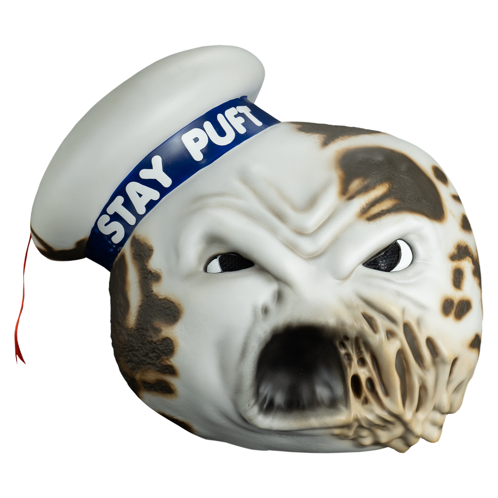 Mask, front view.  round white head furrowed brow, large eyes with large black pupils, mouth open, left side of mouth melting.  Charred spots on both sides of the head and the white hat cocked to the side on the right side of the head, blue band with white text reads Stay Puft, red ribbon tassel hanging from the top center.