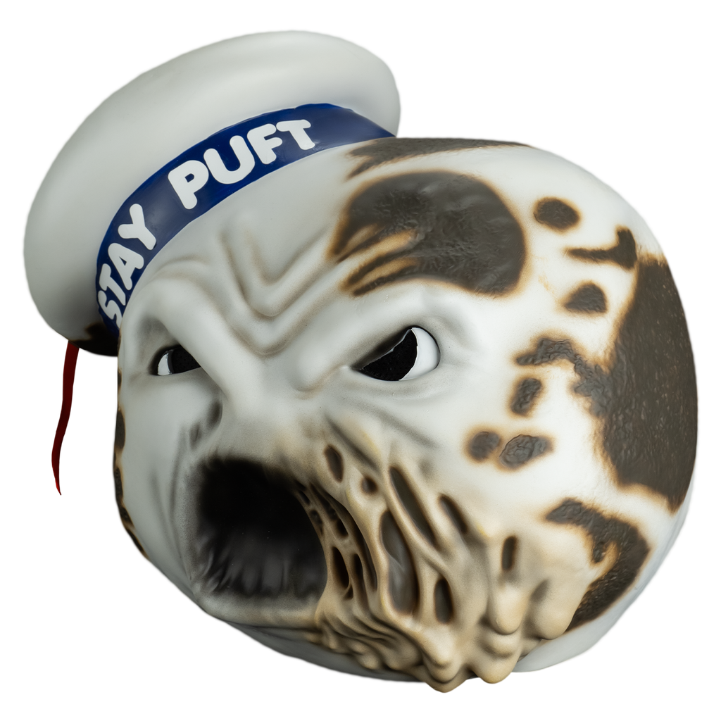 Mask, left view.  round white head furrowed brow, large eyes with large black pupils, mouth open, left side of mouth melting.  Charred spots on both sides of the head and the white hat cocked to the side on the right side of the head, blue band with white text reads Stay Puft, red ribbon tassel hanging from the top center.