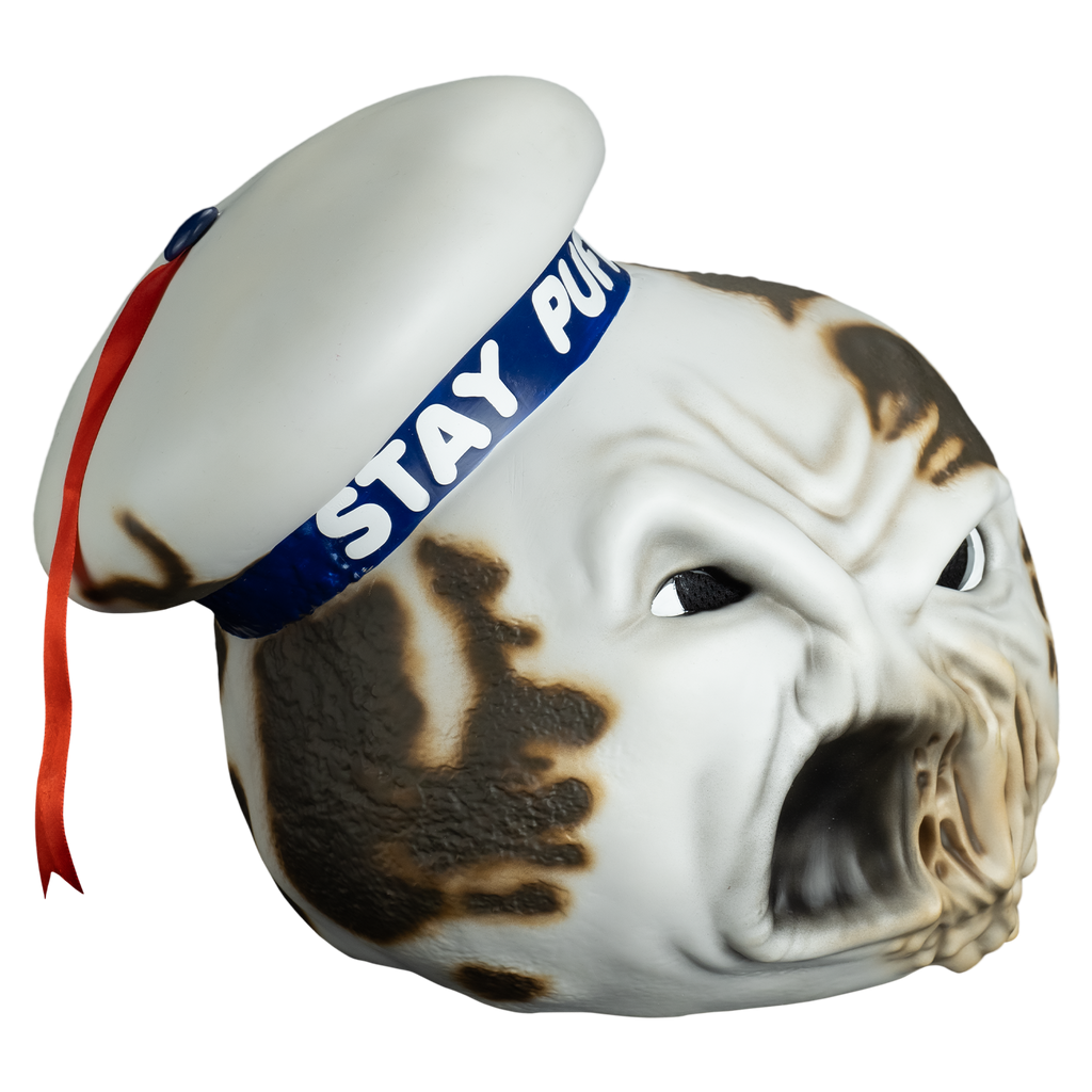 Mask, right view.  round white head furrowed brow, large eyes with large black pupils, mouth open, left side of mouth melting.  Charred spots on both sides of the head and the white hat cocked to the side on the right side of the head, blue band with white text reads Stay Puft, red ribbon tassel hanging from the top center.